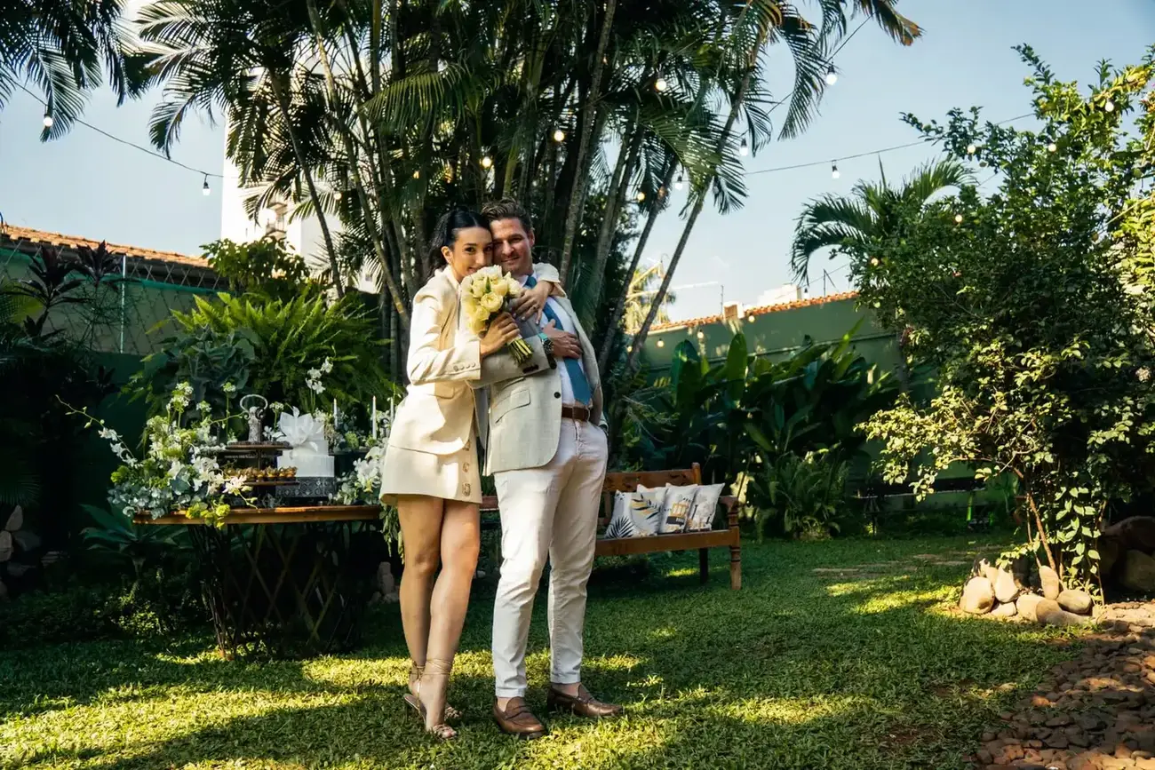 Gabi e Massimo abraçados no jardim decorado do casamento, entre palmeiras e luzes de varal